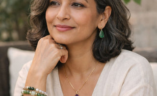 A calm, midlife woman with salt-and-pepper hair wearing a neutral linen top, styled with Stone Story jewelry including a purple stone amethyst pendant, green drop earrings, and stacked beaded bracelets.
