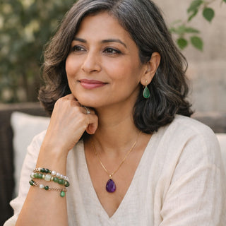 A calm, midlife woman with salt-and-pepper hair wearing a neutral linen top, styled with Stone Story jewelry including a purple stone amethyst pendant, green drop earrings, and stacked beaded bracelets.