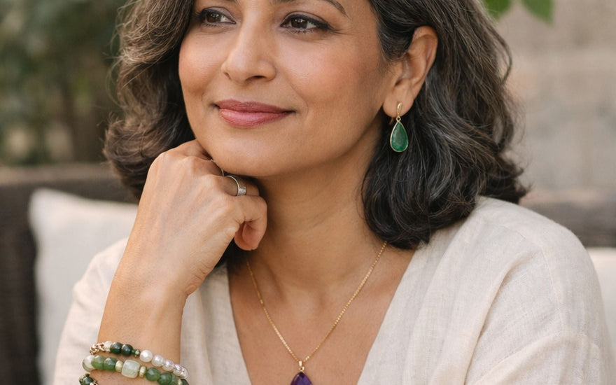 A calm, midlife woman with salt-and-pepper hair wearing a neutral linen top, styled with Stone Story jewelry including a purple stone amethyst pendant, green drop earrings, and stacked beaded bracelets.