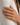 A close-up of a hand wearing three slim rings on the ring finger, set against a soft white blazer. The rings feature gold, silver, and rose gold bands with pavé-set clear stones.
