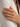 A close-up of a hand wearing three slim rings on the ring finger, set against a soft white blazer. The rings feature gold, silver, and rose gold bands with pavé-set clear stones.